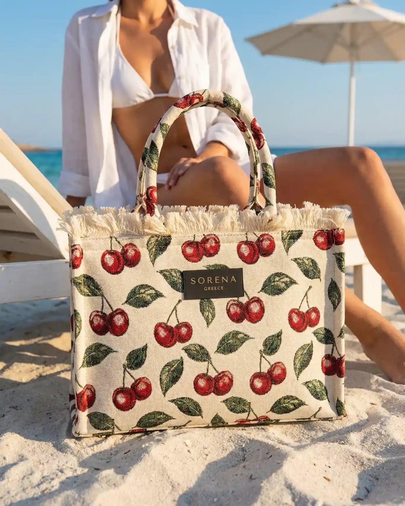 Sorena Greece Cherry Medium Tote Bag im Sand am Meer, Frontansicht, Jacquard Canvas Kirschrot handgefertigter Sommershopper Griechenland Produktpräsentation.