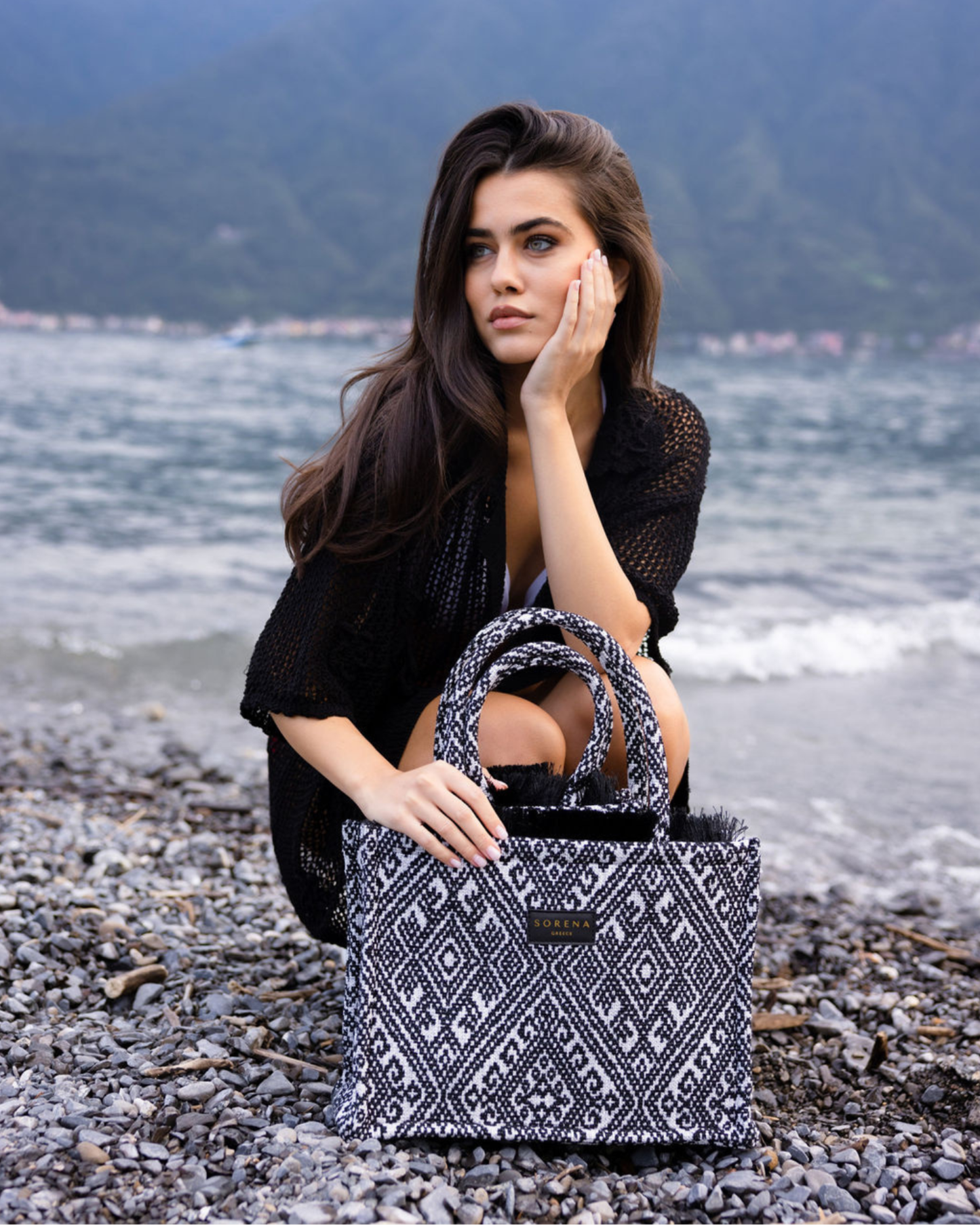 Model sitzt auf Felsen und hält die handgemachte SORENA ROXANI Shopper – vegane, nachhaltige Tasche mit kontrastreichem Grafikmuster in Schwarz-Weiß, ideal für City-Dates und Business-Looks.